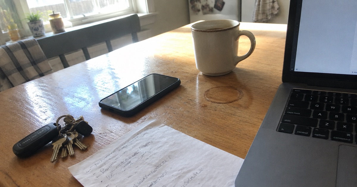 Phone snapshot of a kitchen table on a weekday morning: a phone, a laptop, a scribbled list, keys and a mug.