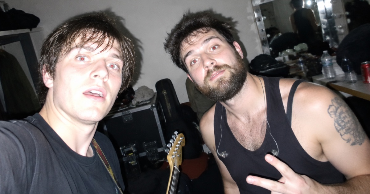 Flash-lit phone snapshot of two musicians from different bands crammed together backstage in a cramped dressing room.
