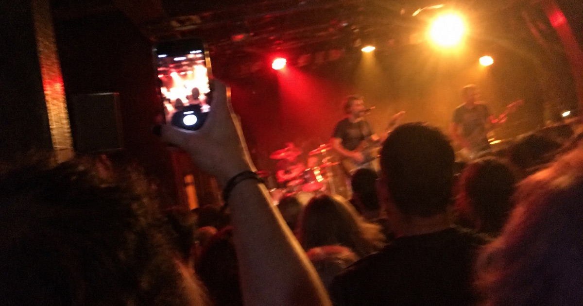 Phone snapshot from within a crowd, a hand raised holding a glowing phone toward a blurred stage.