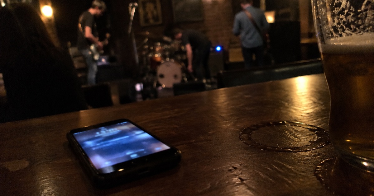 Phone snapshot of a phone face up on a scratched bar top beside a pint glass, band setting up out of focus behind.