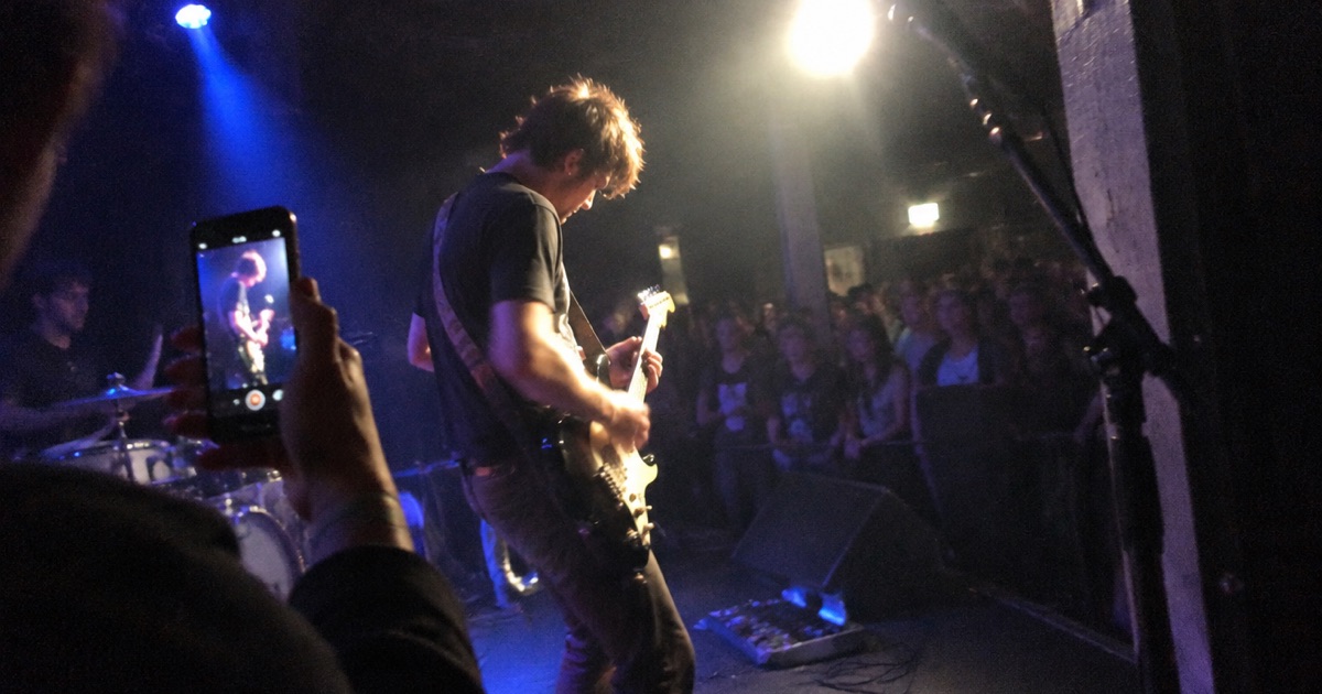 Phone snapshot from the wings of a club stage, a bandmate filming the guitarist vertically on a phone.