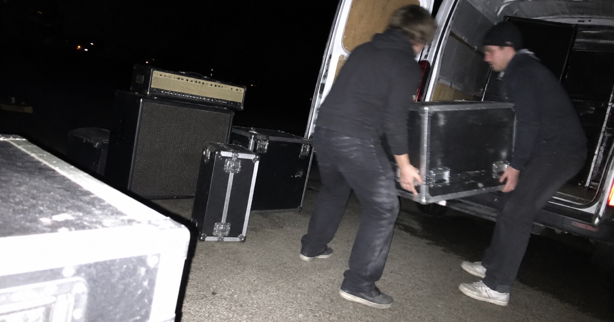 Flash-lit phone snapshot of load-out behind a venue at night, amps and cases by an open van door.