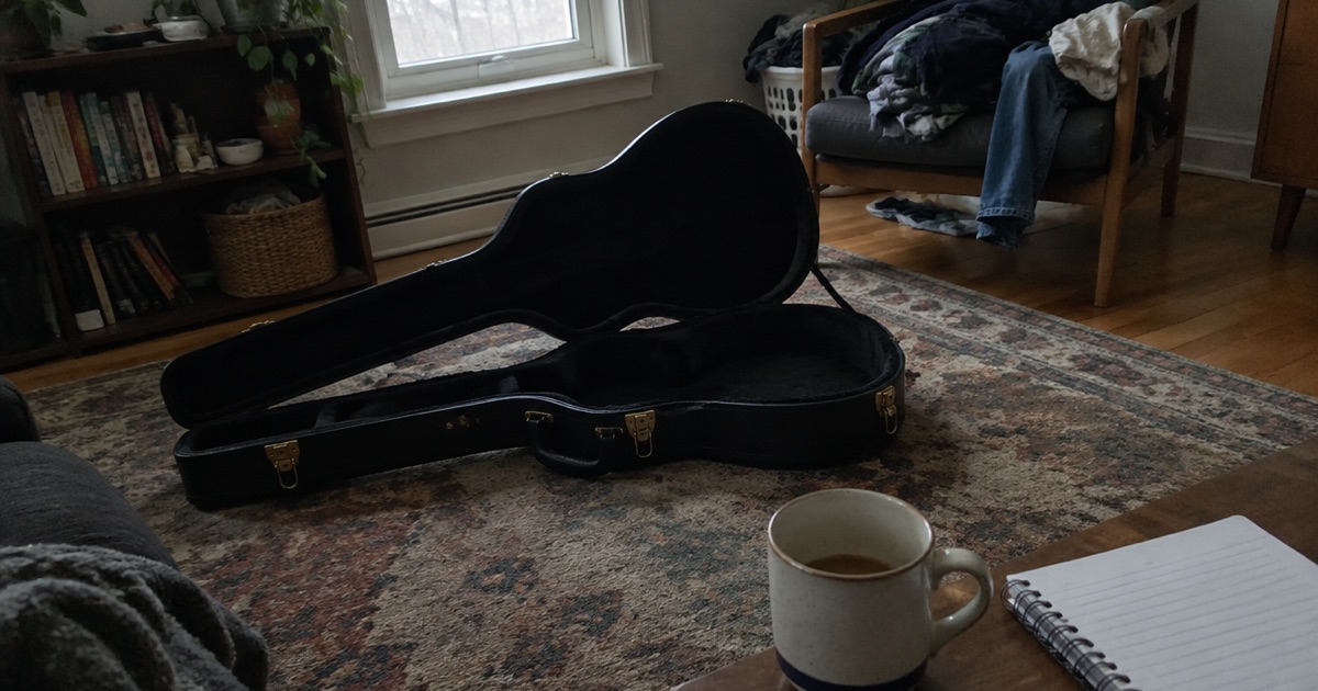 Phone snapshot of a guitar case open on a living room rug with a coffee mug and a notebook beside it.